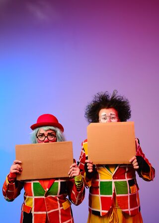 Two clowns a man and a woman with makeup in bright colored costumes are holding a blank cardboard for inscription. April Fools Day concept. Circus performance with a clown.の写真素材