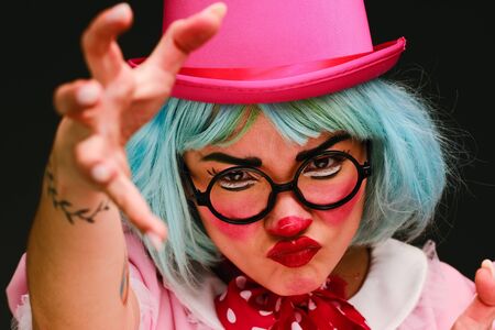 Close-up portrait of a clown girl with makeup, blue hair, in a pink hat, pink dress and glasses. A clown shows different human emotions. April Fools Day concept. Circus performance with a clown.の写真素材