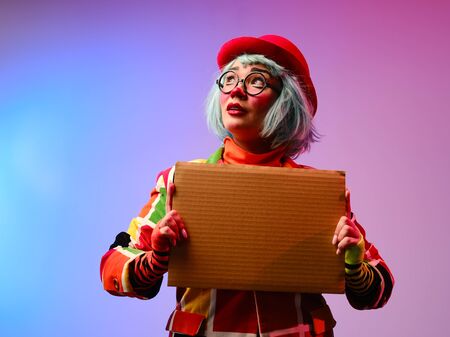 A clown girl with makeup, blue hair, in a red hat, a colored checkered jacket and glasses holds in her hands a blank cardboard for inscription. April Fools Day concept.の写真素材
