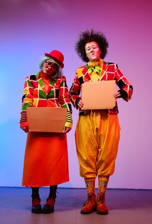 Two clowns a man and a woman with makeup in bright colored costumes are holding a blank cardboard for inscription. April Fools Day concept. Circus performance with a clown.の写真素材