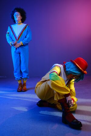 Two clowns a man and a woman with makeup in colored tracksuits in the style of the 80s or 90s posing against a trendy neon background.Party disco 80s 90s vibes.Creative art purple color styleの写真素材