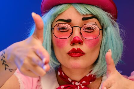 Close-up portrait of a clown girl with makeup, blue hair, in a pink hat, pink dress and glasses. A clown shows different human emotions. April Fools Day concept. Circus performance with a clown.の写真素材