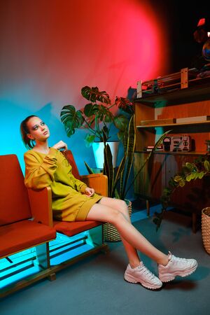 A young attractive girl with bright makeup and in colored clothes sits on a red armchair against a fashionable neon background. The interior of a modern teenager - a lot of flowers and longboardsの写真素材