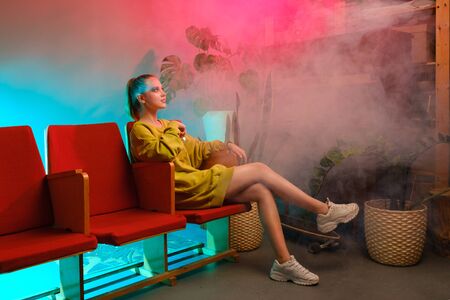 A young attractive girl with bright makeup and in colored clothes sits on a red armchair against a fashionable neon background. The interior of a modern teenager - a lot of flowers and longboardsの写真素材