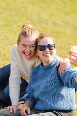 Young girls sit on the grass in the park and take a selfie on a smartphone. Girls enjoy the weekend outdoors. Active rest, health supportの写真素材