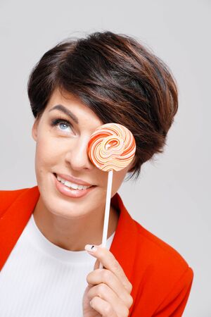 Studio portrait of woman with short dark hair in a red jacket holding colorful lollipop against plain backgroundの写真素材