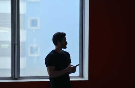 Portrait of a young stylish man with a beard and dark hair in a black T-shirt stands near the window and uses a smartphone.の写真素材