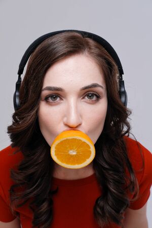 Studio portrait of young woman with headphones and orange covering mouth on plain backgroundの写真素材