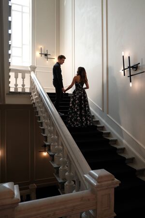 Young beautiful couple - a girl in a magnificent dress and a guy in a black shirt and pants stand or sit on a white staircase inside a large beautiful building. Concept of romance and loveの写真素材