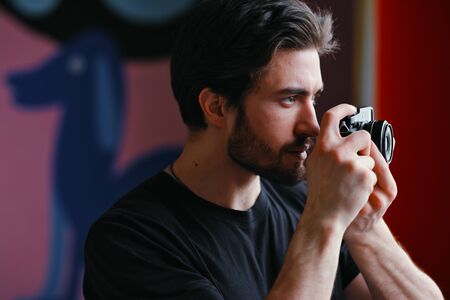 Close-up portrait of a young stylish man with a beard and dark hair in a black T-shirt. Male photographer uses a mirrorless camera to take a picture.の写真素材