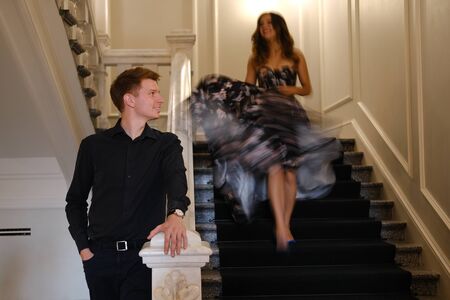 Young beautiful couple - a girl in a magnificent dress and a guy in a black shirt and pants stand or sit on a white staircase inside a large beautiful building. Concept of romance and loveの写真素材