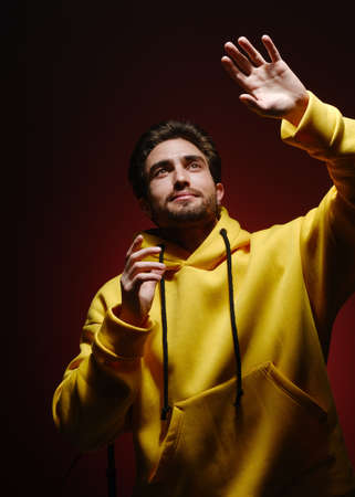 A young man of 25-30 years old in a yellow sweatshirt emotionally pulls his hands up on red wall...