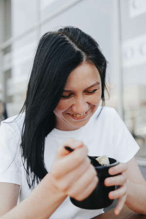 A young woman with black hair in a white T-shirt sits on an open veranda in a cafe and eats food from disposable plastic dishes. Street food, the new reality after the pandemicの写真素材