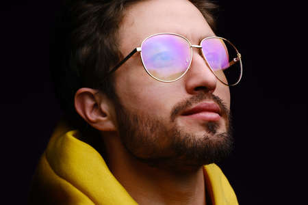 A young man of 25-30 years in glasses and a yellow sweatshirt emotionally poses on a black background. The concept of modern fashion photography. Brutal man portraitの写真素材