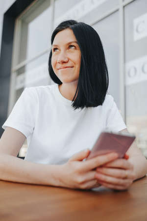 A young woman with black hair in a white T-shirt sits on an open veranda in a cafe and uses a smartphone while waiting for her order and takeaway.の写真素材