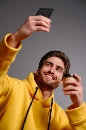 Young smiling guy in a yellow sweatshirt with a disposable paper glass of coffee in hand makes selfie on a smartphone on a gray backgroundの写真素材