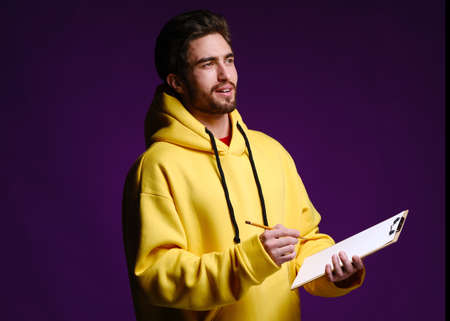 A young man of 25-30 years in glasses and a yellow sweatshirt holding a clipboard with clip in one hand and a pencil in the other on a purple background. White mockup blank. Office conceptの写真素材