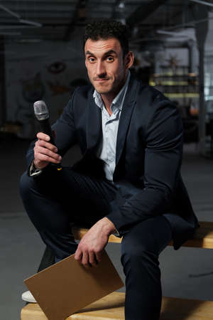 Close up portrait of a man 30-34 years old in a white shirt and a dark blue jacket sitting on a chair on a dark background. A man holds a microphone and clipboard with a clip in his hands and holds an event.の写真素材