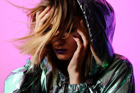 Beautiful girl with bright makeup in a metallic color windbreaker posing on a trendy pink neon...