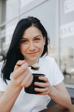 A young woman with black hair in a white T-shirt sits on an open veranda in a cafe and eats food from disposable plastic dishes. Street food, the new reality after the pandemicの写真素材