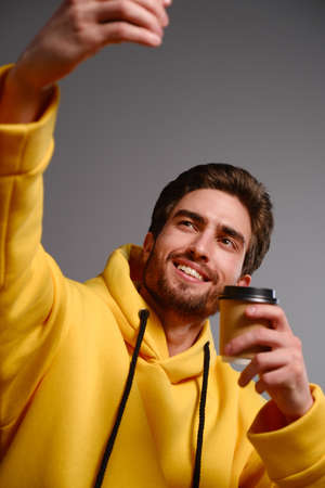 Young smiling guy in a yellow sweatshirt with a disposable paper glass of coffee in hand makes selfie on a smartphone on a gray backgroundの写真素材