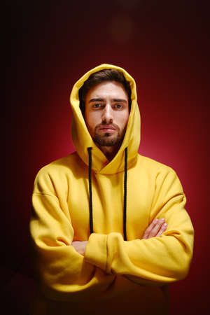 A young man of 25-30 years old in a yellow sweatshirt is standing against a red wall. The concept of modern fashion photography.の写真素材