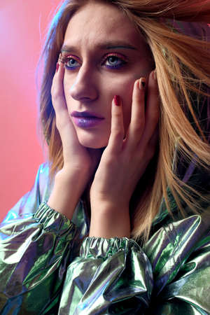 Beautiful girl with bright makeup in a metallic color windbreaker posing on a trendy pink neon...