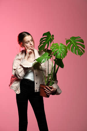 Portrait of a young beautiful girl 20-25 years old with green eyes and long hair. A girl in a pink jacket in black jeans holds in her hands a monstera flower in a pot posing on a pink background.の写真素材