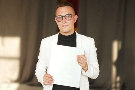 A young man of 30-35 years old in a white jacket, black turtleneck, glasses and a short haircut holds an empty sign in his hands. Template for design, mockup.の写真素材