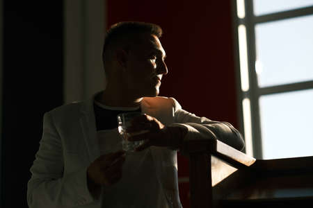 A young man of 30-35 years old in a white jacket, glasses and a short haircut holds in his hands a glass cloth with water. A man stands in the building in the shade.の写真素材