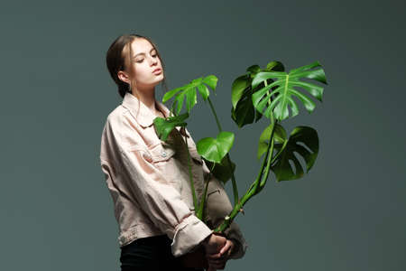 Portrait of a young beautiful girl 20-25 years old with green eyes and long hair. A girl in a pink jacket in black jeans holds in her hands a monstera flower in a pot posing on a gray background.の写真素材
