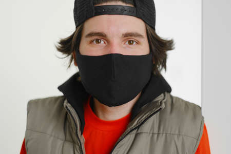 A young man of 25-30 years old in a black protective mask, a cap and a red jacket posing on a gray...