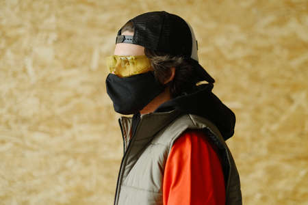 A young man of 25-30 years old in a black protective mask, yellow goggles, a cap and a red jacket posing on a gray background. Coronavirus protection.の写真素材