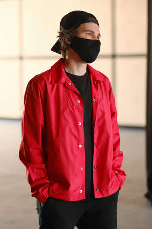 Portrait of a young man 25-30 years old in a black protective mask, black cap and red jacket. Young fashionable man posing over bright background. Fashion photography conceptの写真素材