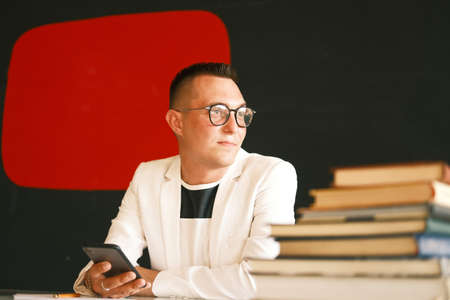 A young school teacher, a man in a white jacket and glasses uses a smartphone and is preparing for a new lesson. The concept of education, school, college and university.の写真素材