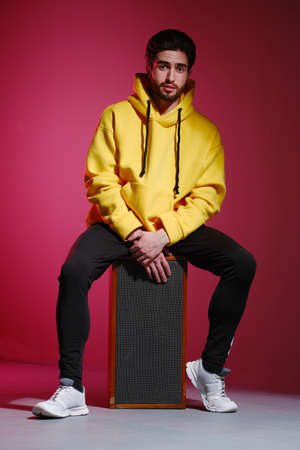 A young man of 25-30 years old in a yellow sweatshirt sitting on an old music column on pink wall background and showing different emotion. The concept of modern fashion photography.の写真素材