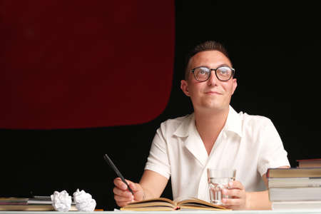 A 30-35-year-old millennial man with a short haircut, wearing glasses, a light shirt, sits at a table and comes up with a script for his vlog. vlogging live feeds on social media networkの写真素材