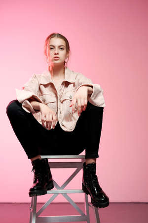 Portrait of a young beautiful girl 20-25 years old with green eyes and long hair. A girl in a pink jeans in black jeans sits on a white wooden bar stool and poses on a pink background.の写真素材