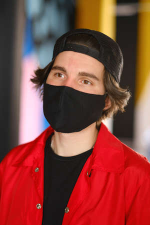 Portrait of a young man 25-30 years old in a black protective mask, black cap and red jacket. Young fashionable man posing over bright background. Fashion photography conceptの写真素材
