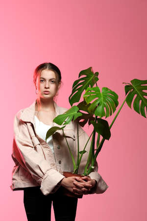 Portrait of a young beautiful girl 20-25 years old with green eyes and long hair. A girl in a pink jacket in black jeans holds in her hands a monstera flower in a pot posing on a pink background.の写真素材