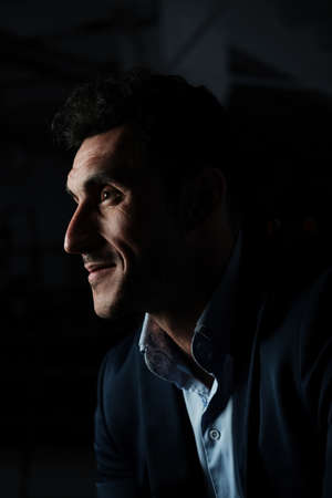 Close up portrait of a serious man 30-34 years old in a blue shirt and a dark jacket on a black background. A man with curly hair, big eyes and stubble. Portrait in the Dark, Low-key lightingの写真素材
