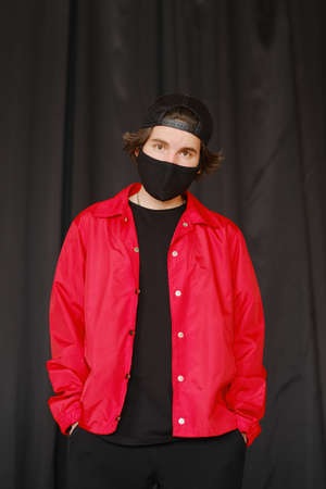 Portrait of a young man 25-30 years old in a black protective mask, black cap and red jacket. Young fashionable man posing over bright background. Fashion photography conceptの写真素材