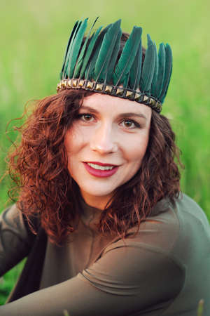 Close-up portrait of a young woman of 25-30 years old with curly red hair, in a green dress and a tiara with feathers. Portrait of a woman on a background of green grassの写真素材