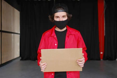 A young man of 25-30 years old in a black protective mask, yellow glasses, a cap and a red jacket holds an empty cardboard sign in his hands. Protest concept. Blank mock up for design, mockupの写真素材