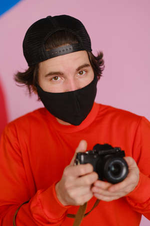 A young man of 25-30 years old in a black protective mask and in a red sweatshirt holds a mirrorless camera in his hands. Male photographer uses a mirrorless camera to take a picture.の写真素材