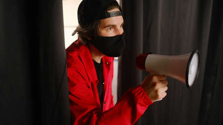 Emotional young man 25-30 years old in a black protective mask, a cap and a red jacket holding a megaphone against a dark background. Protest conceptの写真素材