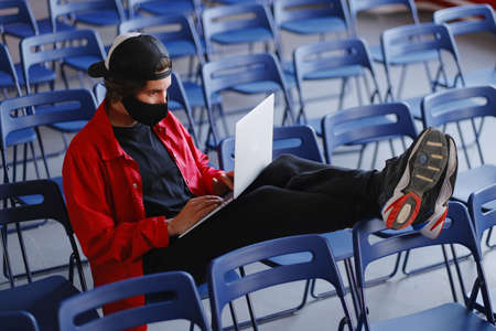 Portrait of a man 25-30 years old in a black protective mask, cap and red jacket. A man sits alone in a large room with many empty blue chairs and works on his laptop. Freelance and lockdown conceptの写真素材
