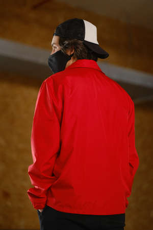 Portrait of a young man 25-30 years old in a black protective mask, black cap and red jacket. Young fashion man posing with his back against a bright background. Clothing for mockup. Fashion photography conceptの写真素材