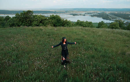 A young woman of 25-30 years old with curly red hair, in a green dress and in a diadem with feathers is standing against the background of beautiful nature at dawn. Solitude with nature. の写真素材