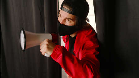 Emotional young man 25-30 years old in a black protective mask, a cap and a red jacket holding a megaphone against a dark background. Protest conceptの写真素材